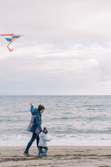 Mom with a kite on a string walks with a little girl by the hand along the seashore