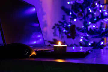 A burning candle and a laptop against the background of a New Year tree. Blackout due to the war in Ukraine
