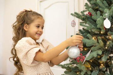 Merry Christmas and Happy Holidays. Cute little child girl in dress is decorating the Christmas tree indoors.