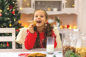 Happy joyful cute little girl in red sweater drinking milk and eating delicious cookie near...