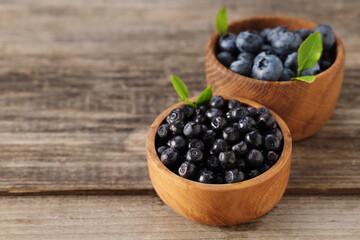 Ripe bilberries and leaves in bowls on wooden table, closeup. Space for text