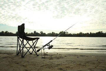Fototapeta na wymiar Folding chair and fishing rod on sand near river, space for text