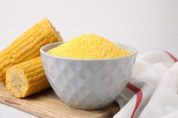 Raw cornmeal in bowl and corn cobs on white table, closeup