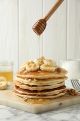 Pouring honey from dipper onto delicious pancakes with bananas and walnuts at white marble table