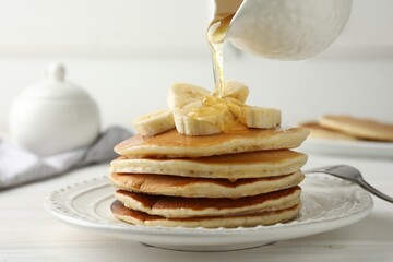 Pouring honey from jug onto delicious pancakes with bananas at white wooden table, closeup