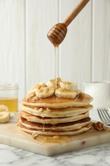 Pouring honey from dipper onto delicious pancakes with bananas and walnuts at white marble table