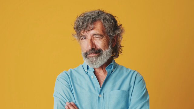 Close-up of an elderly grey-haired bearded man wearing a blue shirt looking camera wink eye blink isolated on orange background in studio