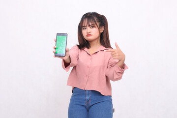 Beautiful happy Asian woman in her 20s wearing casual shirt holding white screen cellphone while frowning pointing at gadget on white background studio portrait for banner ad, banner, billboard