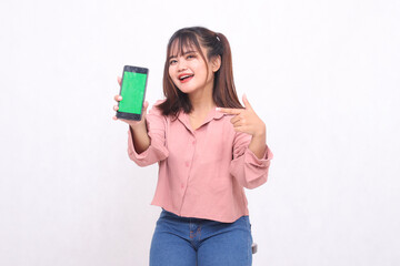 Beautiful happy Asian woman in her 20s wearing casual shirt holding green screen cell phone laughing pointing at gadget on white background studio portrait for banner ad, banner, billboard
