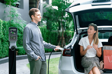 Young couple travel with EV electric car charging in green sustainable city outdoor garden in...