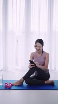 An Asian woman in workout attire is using a smartphone on a yoga mat in her home's sitting room. Ideal for showcasing active lifestyles and a cozy home yoga space