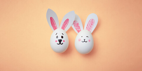 Two white chicken eggs with cute cartoon bunny face and rabbit paper ears on beige background. Preparations for celebration of religious holiday. Happy Easter gift. Copyspace. Trendy peach color 2024.