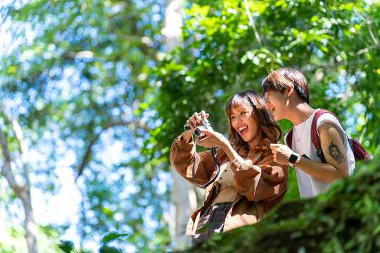 Young Asian man and woman friends travel nature on summer holiday vacation. Generation z people enjoy and fun outdoor lifestyle using camera taking picture of beautiful nature at forest mountain.