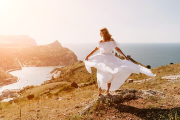 Happy woman in a white dress and hat stands on a rocky cliff above the sea, with the beautiful...