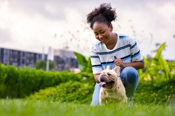 African American woman is playing with her french bulldog puppy while lying down in the grass lawn after having morning exercise in the public park