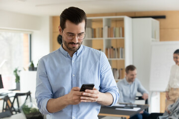 Young Caucasian male employee in office look at smartphone screen browse wireless internet on gadget. Millennial man worker use cellphone text message on device. Technology, communication concept.
