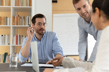 Multiracial businesspeople gather at desk in office brainstorm over business project together. Motivated diverse colleagues talk discuss ideas at meeting or briefing. Teamwork, collaboration concept.