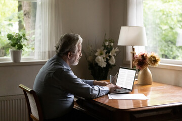 Aged person and tech. Mature grey haired man writer working on book typing text on laptop at cozy home office. Senior elderly male pensioner using pc to write email search information browse websites