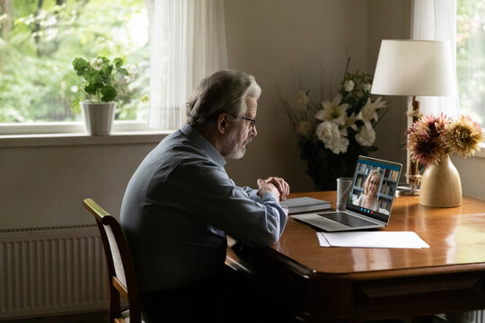 Telecommunication. Older man wearing glasses sit by work desk at home talk with wife by videocall via messenger app on laptop. Aged male pensioner enjoy virtual conversation with smiling mature female