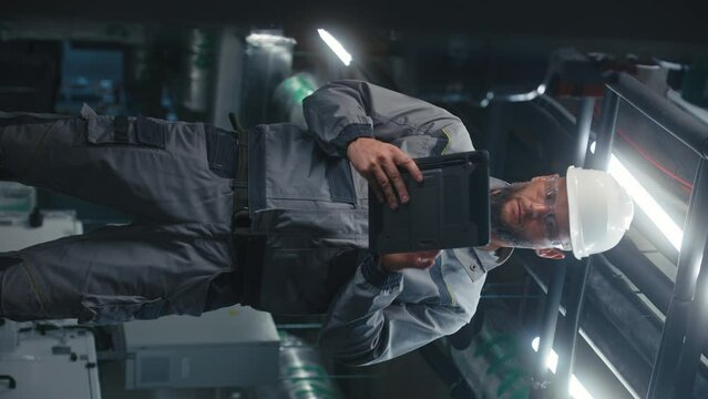 Heavy industry worker wearing protective uniform and hardhat works on factory using digital tablet computer. Professional engineer maintains modern plant or industrial energy facility. Vertical video.