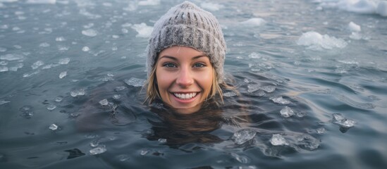 Young woman smiling while swimming in a frozen lake's ice hole, wearing a gray hat and gloves. - obrazy, fototapety, plakaty