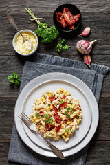 portion of mac and cheese with corn, bacon topped with panko breadcrumbs on plate on dark wooden table with ingredients at background, vertical view from above