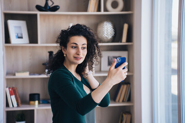 Cheerful woman talks by phone makes video call standing  home against book shelf. Happy female entrepreneur makes selfie at apartment.