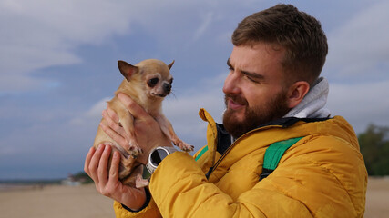Happy young man walking with his little pet Chihuahua or Toy terrier dog on the beach, hold puppy on hands. People love animals concept.