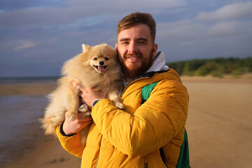 Happy young man walking with his pet Pomeranian Spitz dog on the beach, hold puppy on hands. People love animals concept.