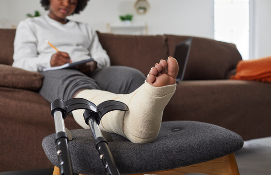 African American woman with broken leg sitting on sofa at home and writing in diary, with her injured foot resting on stool, with black crutches beside. Foot wrapped in bandage closeup. Injury concept