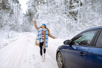 Beautiful young woman near a car in a snowy forest. First snow, great mood.  A traveler poses near a car in nature. Concept of car travel, vacation.