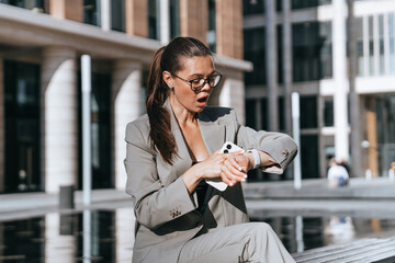Unhappy girl in business suit sitting outside looks at wrist watch with opened mouth, late....