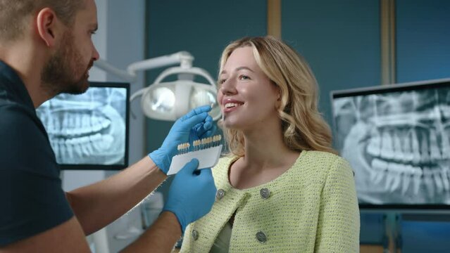 Cute blonde woman standing smiling while young dental expert holding samples of implants choosing suitable shade for veneers. Clinic with modern equipment. High quality 4k footage