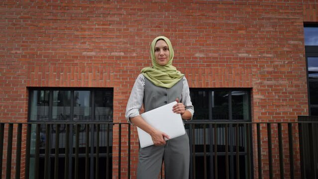 Beautiful woman in hijab standing on city street. Muslim businesswoman with laptop in hands. Iran, Afganistan female teacher in front of school building.