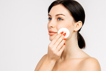 Charismatic, beautiful young woman touching a cotton pad to her face on a light isolated...