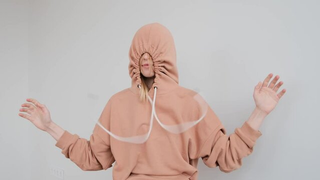 Portrait of a young teenage woman with a large birthmark on her face in a peach beige hoodie tightens the hood with a drawstring, hiding and then revealing knot looking at camera on a white background