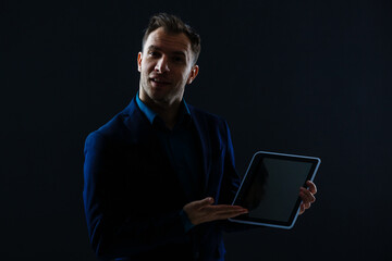 A serious handsome young businessman in a suit using a tablet in front of a dark grey background in a studio.