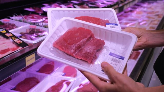 Packaged pieces of raw fresh beef laid out in supermarket refrigerator. Human hands select piece of fresh beef. Concept: you can always find right cut of fresh beef for steak on supermarket shelves