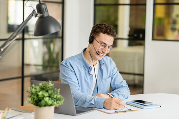 Positive handsome millennial european guy manager in headphones with laptop at table