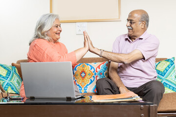 Overjoyed indian senior couple happily living retirement life, senior couple doing high five while sitting on sofa at living room - obrazy, fototapety, plakaty