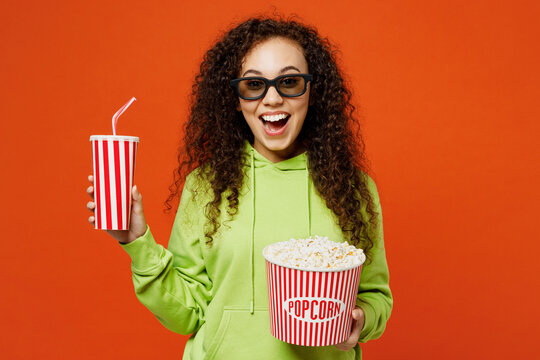 Young excited woman of African American ethnicity she wear green hoody casual clothes 3d glasses watch movie film hold bucket of popcorn cup of soda pop in cinema isolated on plain orange background.