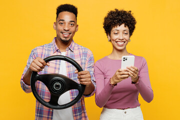 Young couple two friends family man woman of African American ethnicity wear purple casual clothes together hold steering wheel driving car use mobile cell phone isolated on plain yellow background.