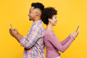 Side view young cool smiling couple two friends family man woman wear purple casual clothes together hold in hand use mobile cell phone stand back to back isolated on plain yellow orange background.