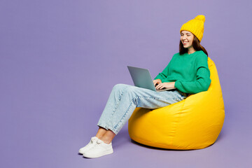Full body young IT woman wear green sweater yellow hat casual clothes sit in bag chair hold use work on laptop pc computer isolated on plain pastel light purple background studio. Lifestyle concept.