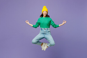 Full body young woman wear green sweater yellow hat casual clothes jump high hold spread hands in yoga om aum gesture relax meditate try to calm down isolated on plain pastel light purple background.