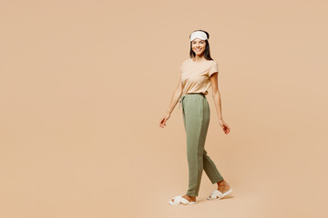 Full body side view smiling young happy calm Latin woman wears pyjamas jam sleep eye mask rest relax at home walk go look camera isolated on plain pastel beige background. Good mood night nap concept.