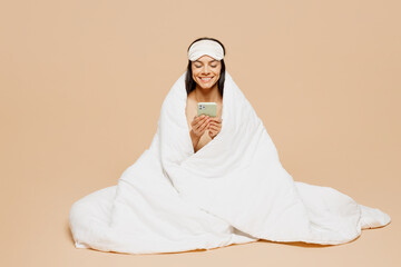 Full body young happy calm Latin woman wrapped in duvet wears jam sleep eye mask rest relax at home...