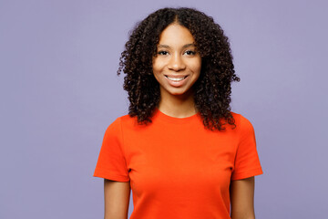 Little smiling cheerful satisfied kid teen girl of African American ethnicity she wear orange t-shirt look camera isolated on plain pastel light purple background studio. Childhood lifestyle concept.