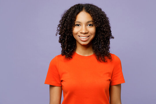 Little Smiling Cheerful Satisfied Kid Teen Girl Of African American Ethnicity She Wear Orange T-shirt Look Camera Isolated On Plain Pastel Light Purple Background Studio. Childhood Lifestyle Concept.