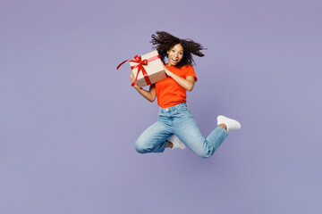 Full body little kid teen girl of African American ethnicity wear orange t-shirt jump high hold present box with gift ribbon bow isolated on plain purple background studio Childhood lifestyle concept